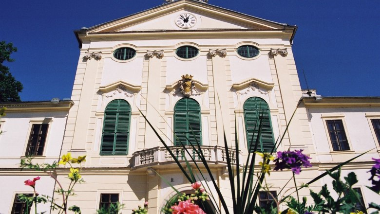 Historisches Gebäude mit grünen Fensterläden und Uhr auf der Fassade, umgeben von bunten Blumen.