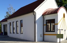 Weiße Kapelle mit rotem Dach und gelben Akzenten, umgeben von gepflastertem Boden und Pflanzen.