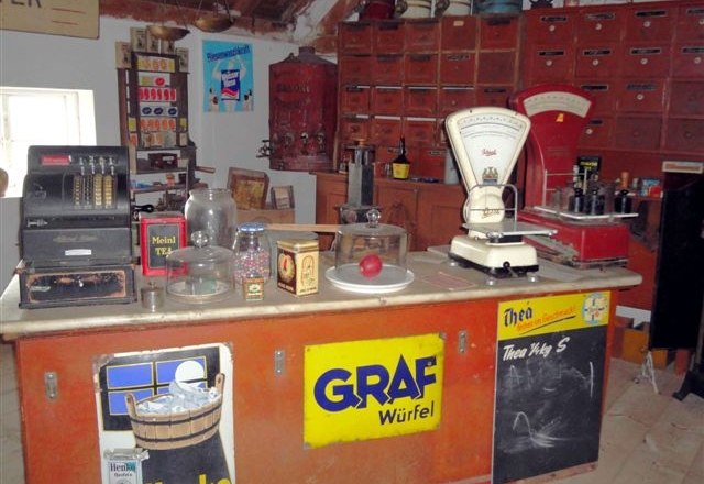 Ein historischer Dorfladen mit alter Registrierkasse, Waage und diversen Produkten auf einem Tresen.