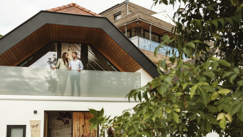 Ein modernes Geb&auml;ude mit einem Paar auf dem Balkon, umgeben von B&auml;umen.