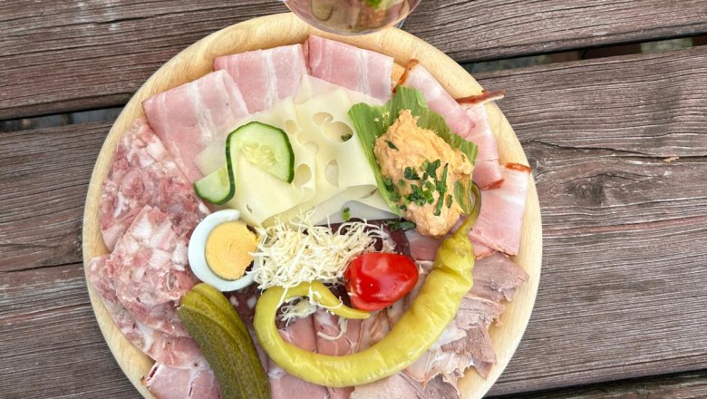 Ein Teller mit Aufschnitt, Käse, Ei, Gurke, Paprika und Brotaufstrich auf einem Holztisch. Daneben ein Glas Roséwein mit Strohhalm.