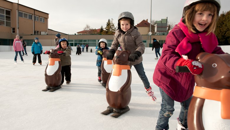 Kinder beim Schlittschuhlaufen mit Pinguin-Hilfen auf einer Eisbahn.