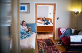 Innenansicht einer Ferienwohnung mit zwei Zimmern. Im Vordergrund sitzt ein M&auml;dchen auf einem Bett, im Hintergrund lesen zwei Personen auf einem anderen Bett.