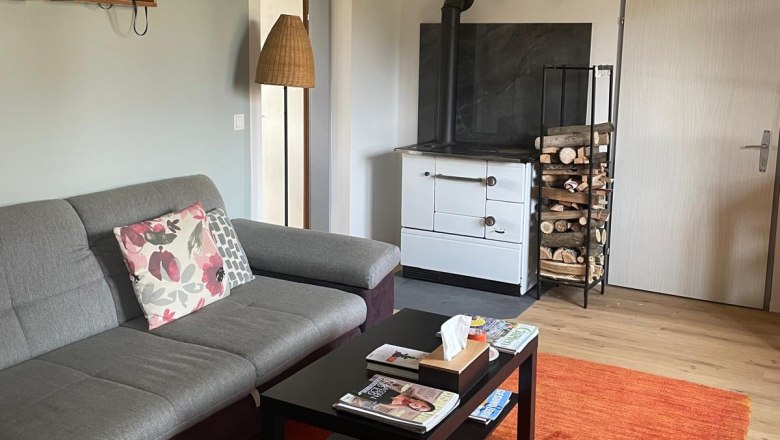 Gem&uuml;tliches Wohnzimmer mit grauem Sofa, Couchtisch, rotem Teppich und Holzofen.