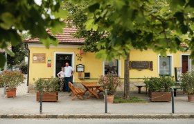 Ein gelbes Gasthaus mit rotem Dach, umgeben von Bäumen. Zwei Personen stehen vor dem Eingang.