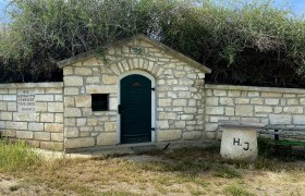 Steinhütte mit grünem Tor und Inschrift, umgeben von Büschen und Gras.