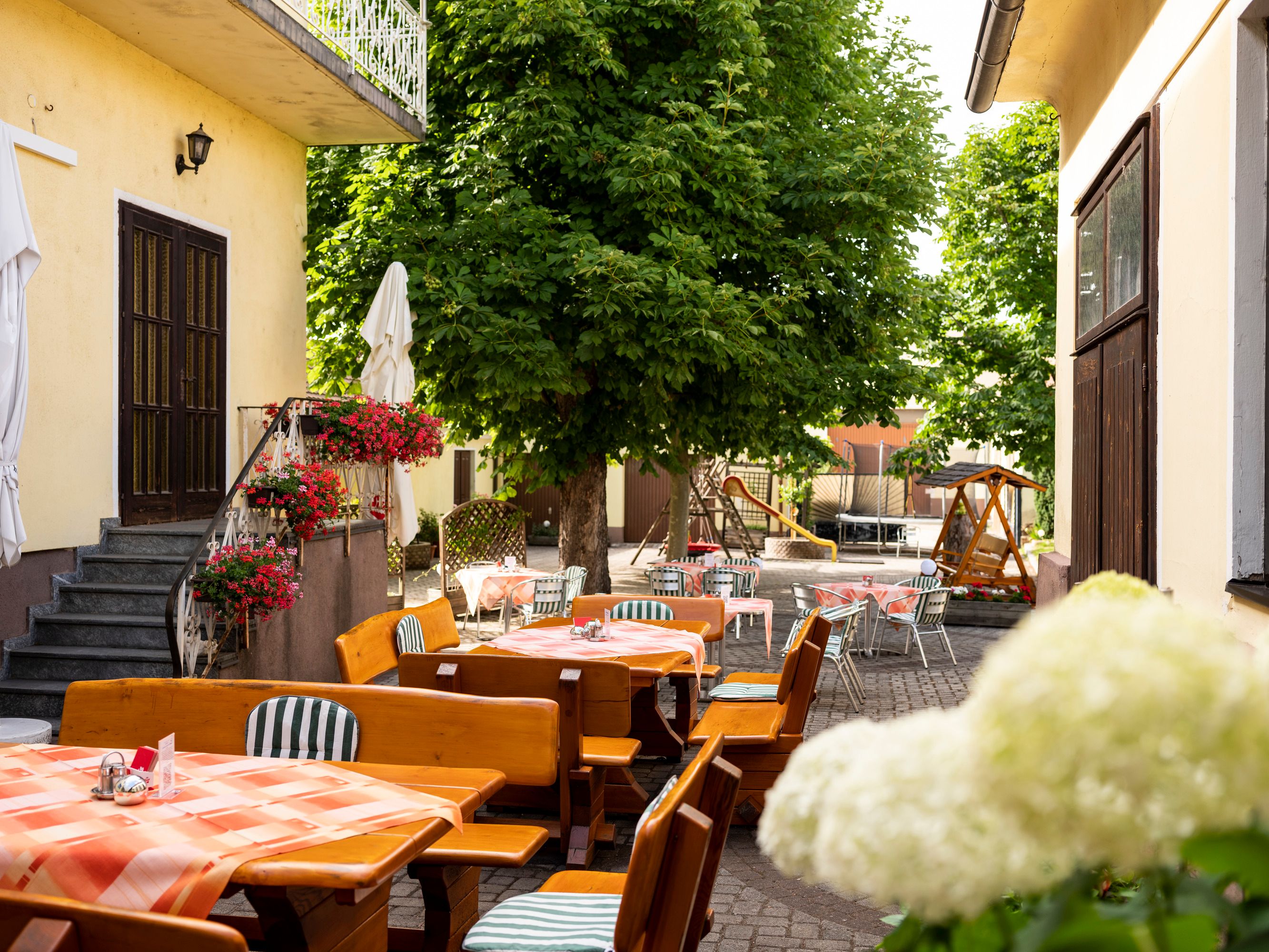 Ein schattiger Gastgarten mit Holztischen und Stühlen, umgeben von Bäumen und Blumen, ideal für heiße Sommertage.