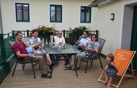Eine Familie sitzt auf einer Terrasse an einem Tisch mit Getränken. Im Hintergrund sind Blumen und ein Gebäude zu sehen.