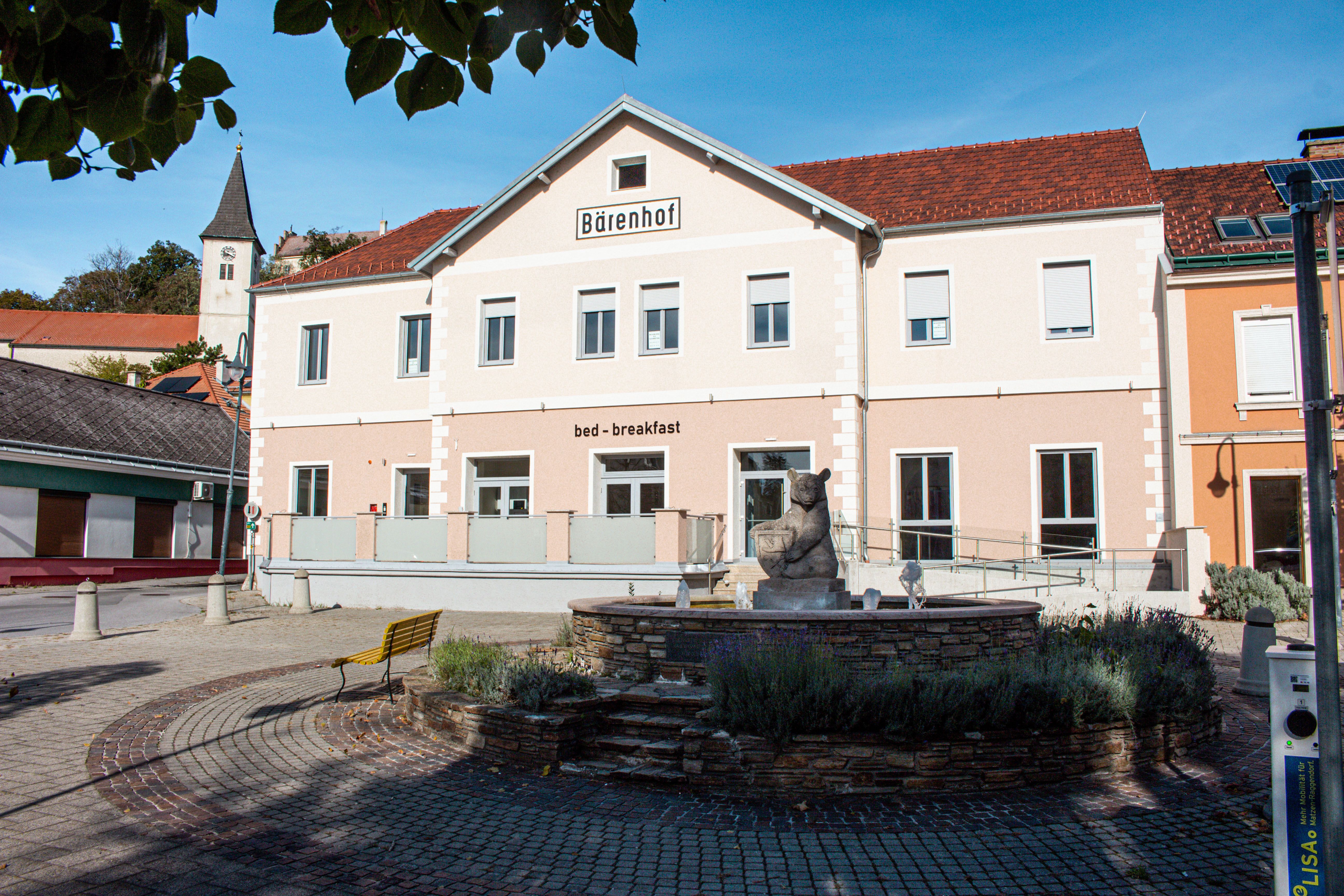 Ein Gebäude mit der Aufschrift 'Bärenhof' und 'bed - breakfast' in einer sonnigen Umgebung mit einem Brunnen im Vordergrund.