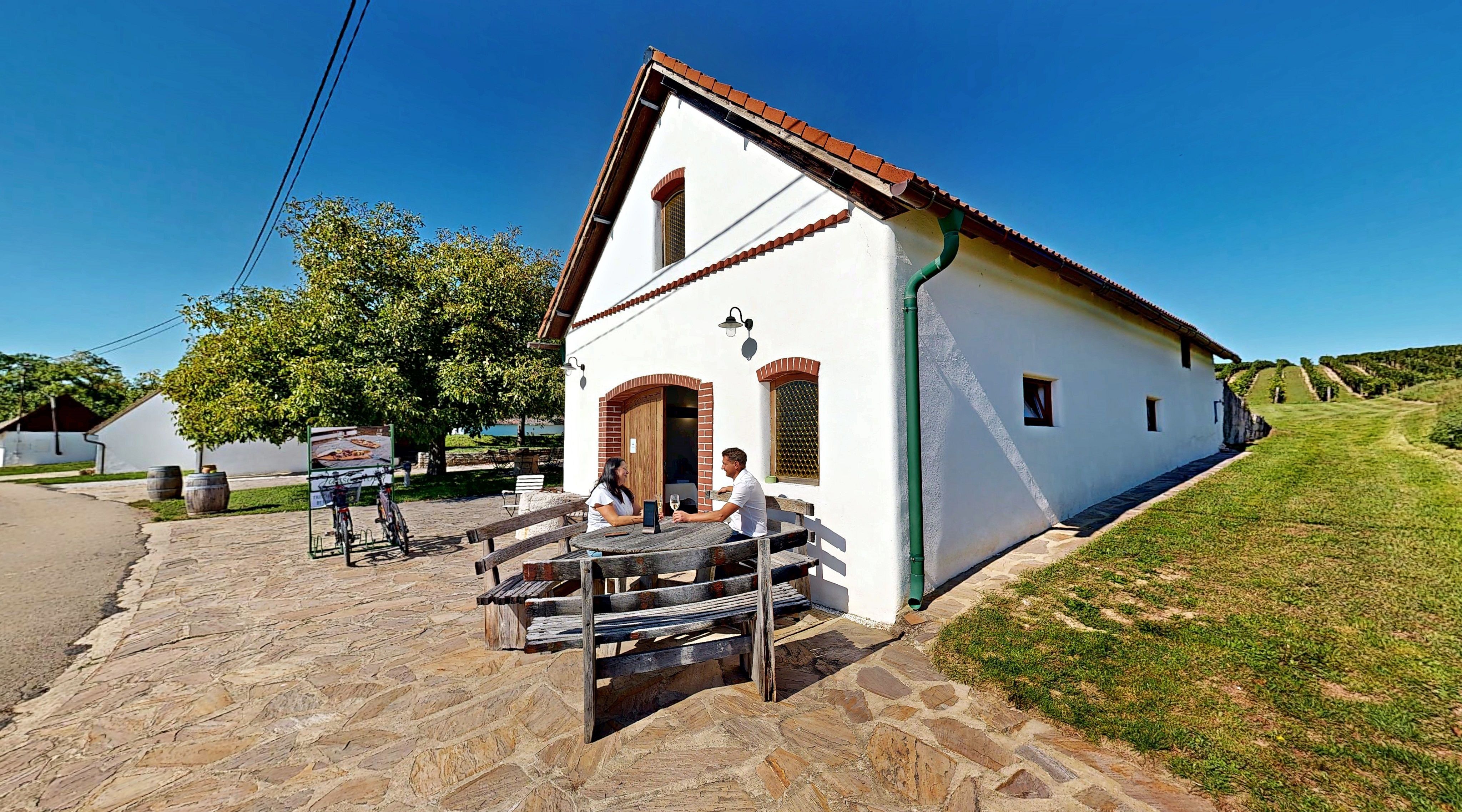 Zwei Personen sitzen an einem Holztisch vor einem weißen Gebäude mit rotem Dach, umgeben von grüner Landschaft und Weinbergen.