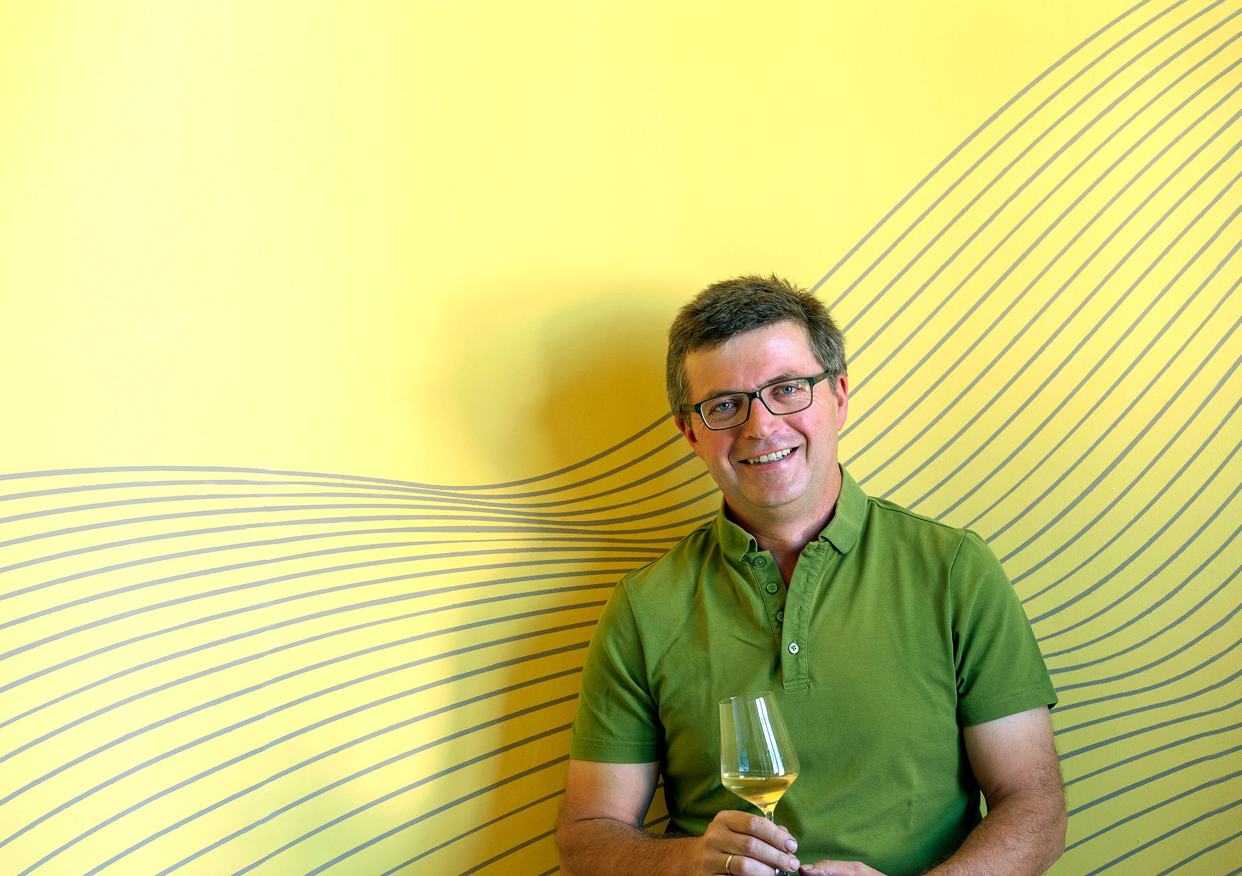 Mann mit Brille und grünem Hemd hält ein Weinglas vor gelbem Hintergrund mit Linienmuster.