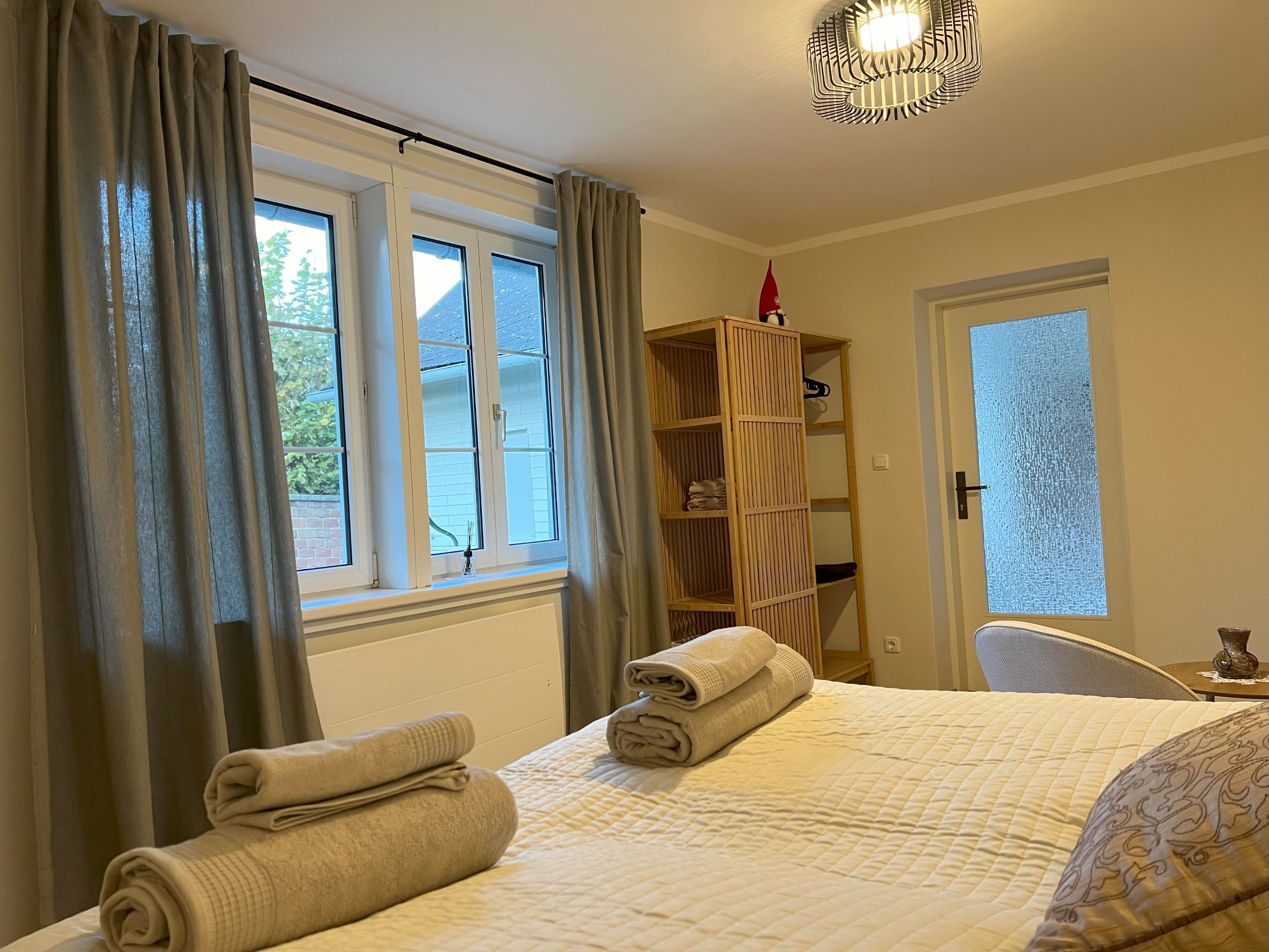 Ein gemütliches Schlafzimmer mit einem Bett, auf dem Handtücher liegen, einem Fenster mit Vorhängen und einem Regal mit Dekoration.