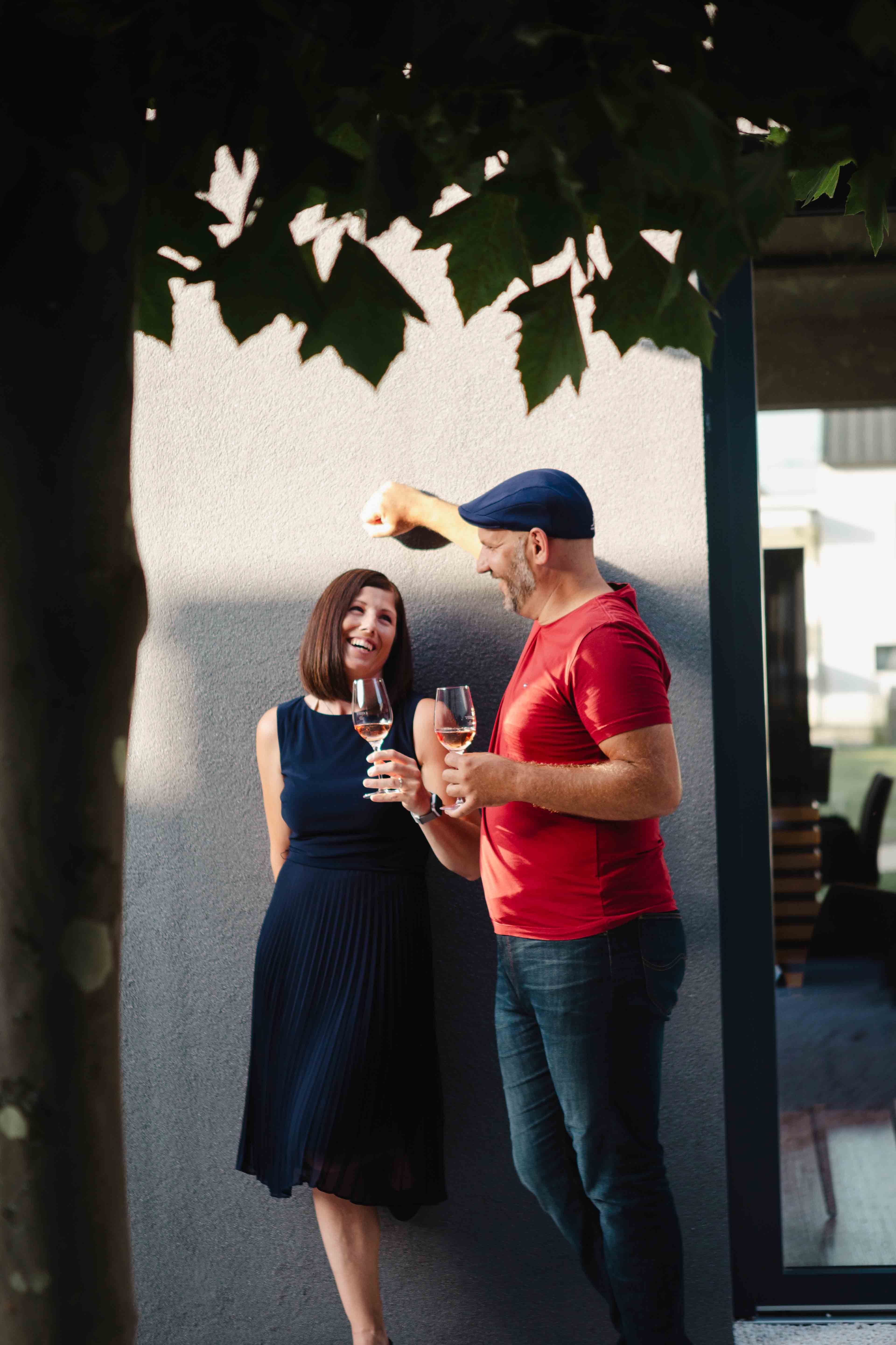A man and a woman stand smiling against a wall, holding glasses of wine and shaded by leaves.