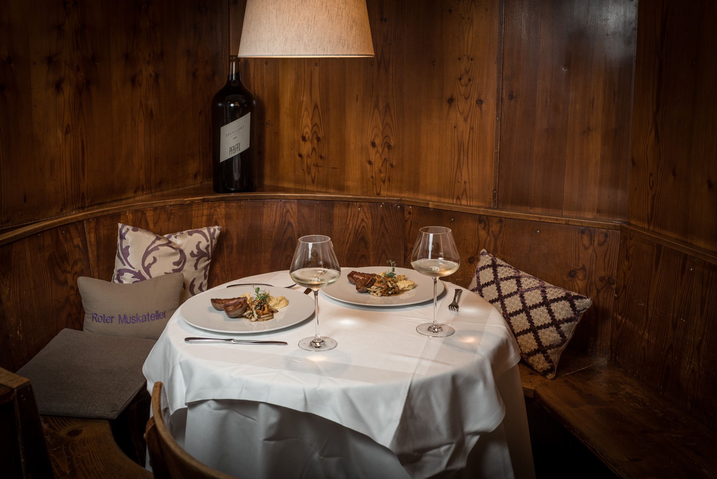 Gemütliche Gaststube mit gedecktem Tisch, zwei Tellern mit Speisen und Weingläsern, Holzwand und Kissen.