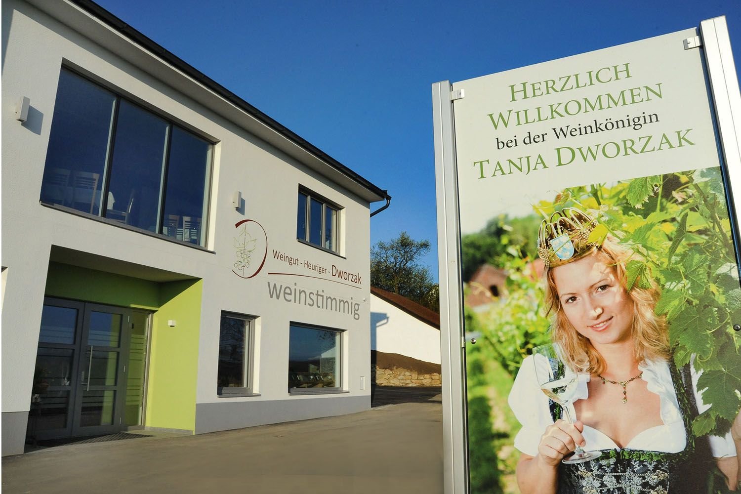 Ein modernes Gebäude mit einem Schild, das eine Frau in Tracht mit einem Weinglas zeigt. Text: Herzlich willkommen bei der Weinkönigin Tanja Dworzak.