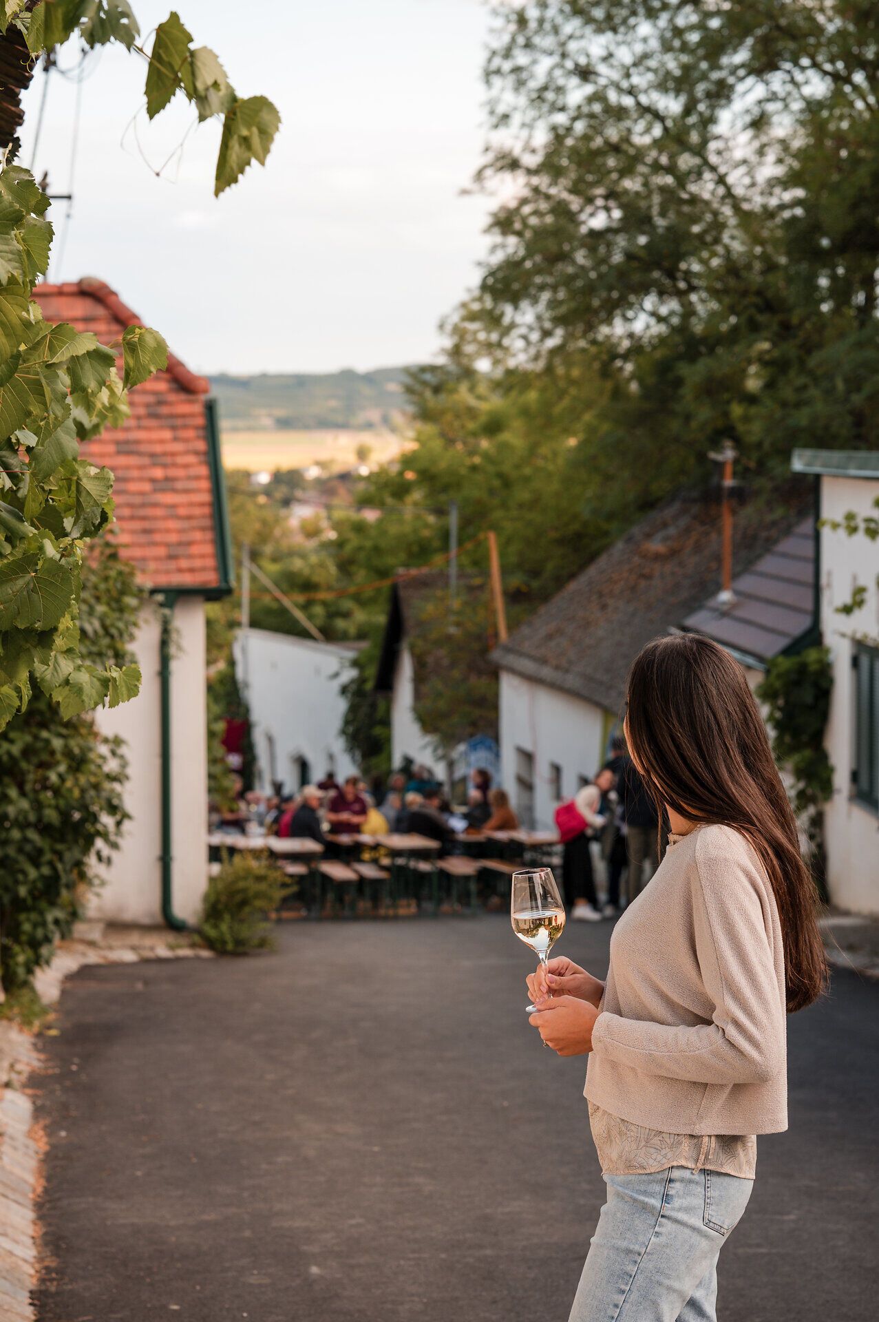 Eine Frau steht mit einem Glas Weißwein in der Hand in einer Kellergasse und betrachtet das gesellige Treiben, das im Rahmen des Kellergassenfestes herrscht.