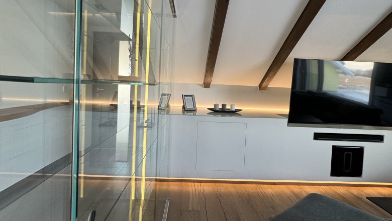 Modernes Wohnzimmer mit Holzboden, Glasvitrine, TV und schr&auml;ger Decke mit Holzbalken.