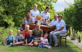 Eine Familie sitzt und steht um einen Tisch im Freien, umgeben von Bäumen und Gras.