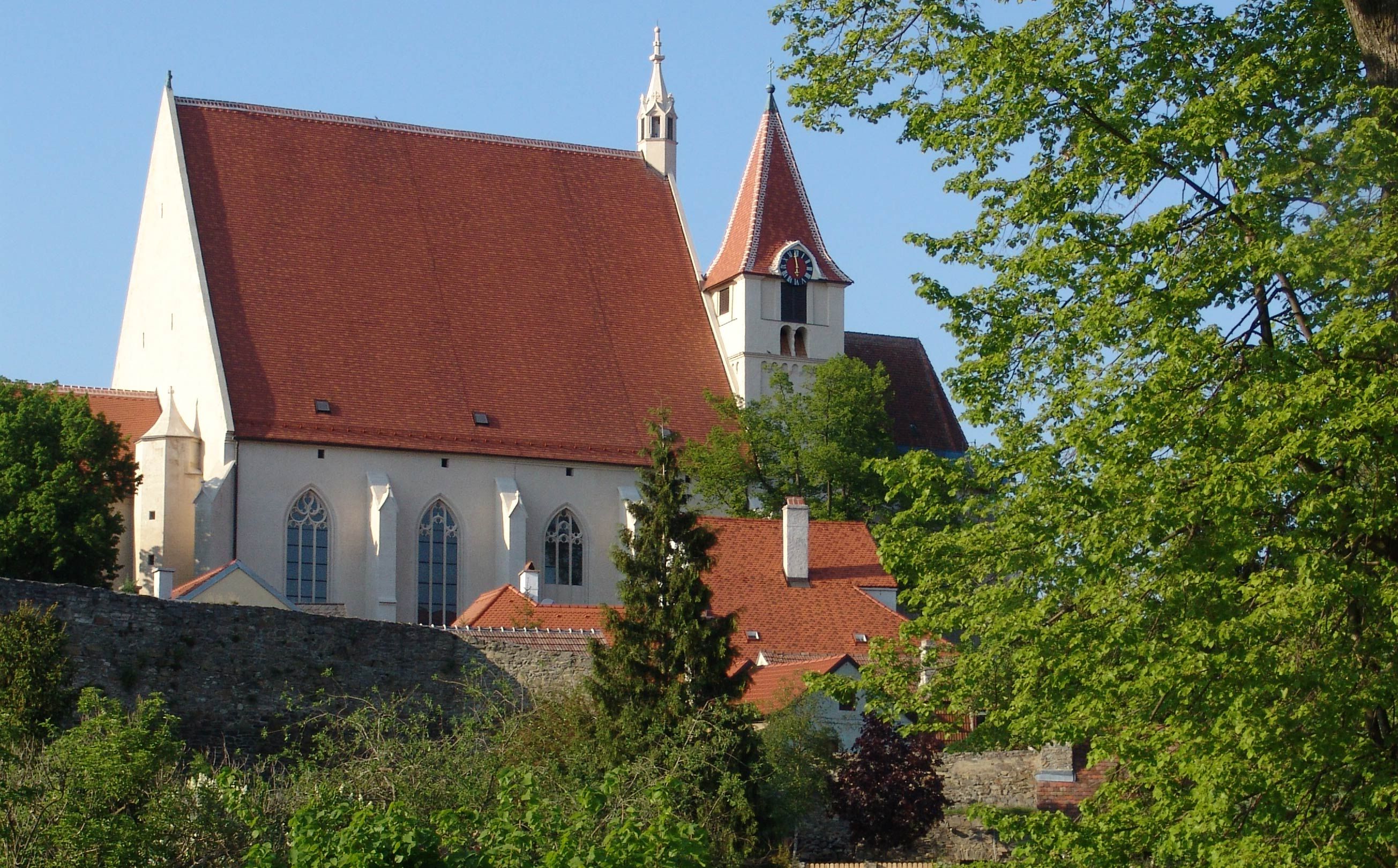 Pfarrkirche St. Stephan mit rotem Dach und Turm, umgeben von Bäumen und einer Steinmauer.