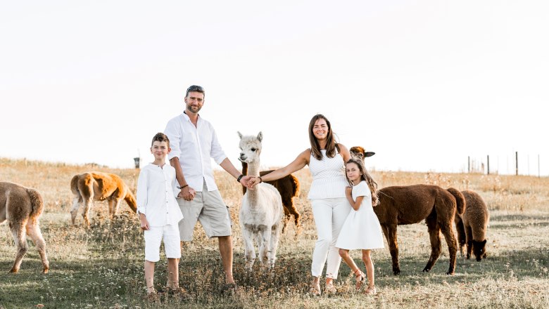 Eine Familie in wei&szlig;er Kleidung steht auf einer Wiese mit Alpakas.