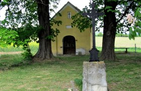 Gelbe Kapelle mit Kreuz und Bäumen im Vordergrund.