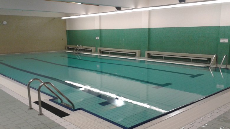 Innenansicht eines leeren Hallenbads mit grün gefliesten Wänden und einem großen Schwimmbecken.