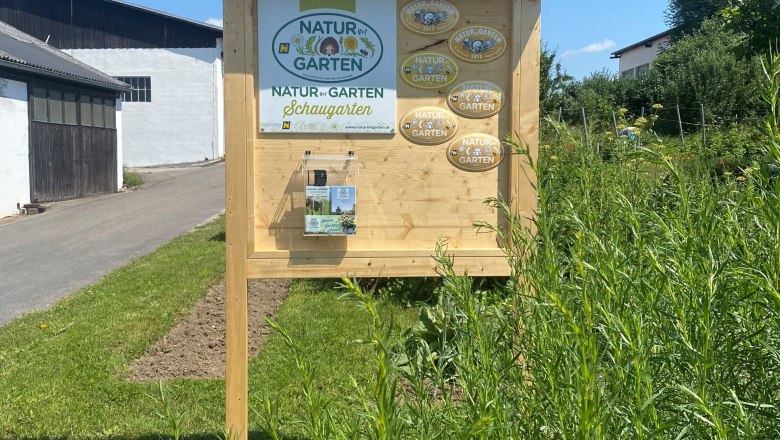 Holztafel mit Natur im Garten Schaugarten-Logo und Plaketten, umgeben von Pflanzen.