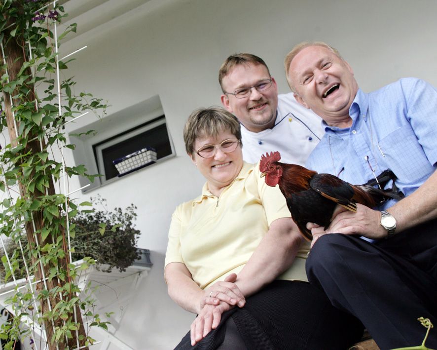 Three laughing people are sitting on a staircase, one is holding a rooster.