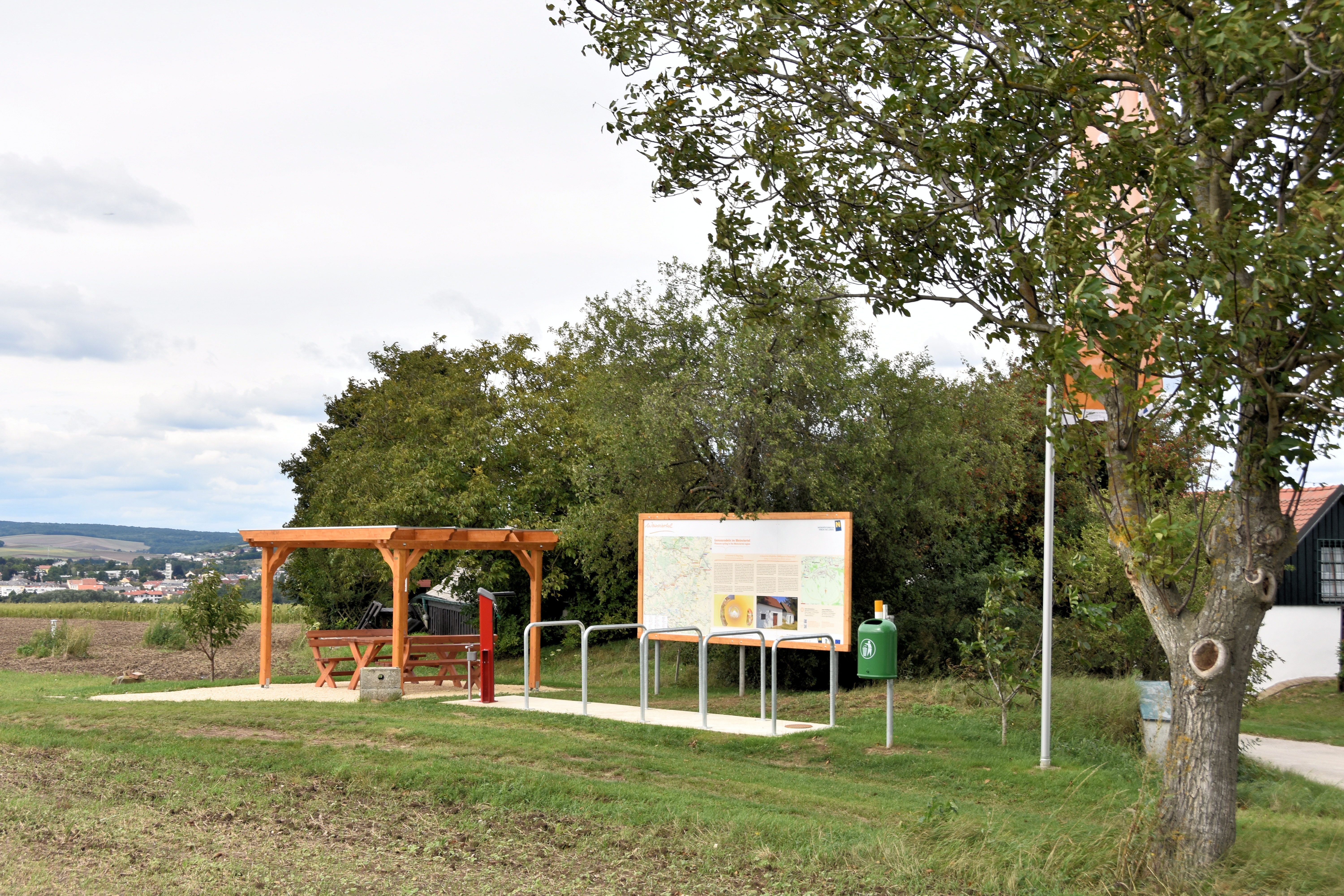 Ein Rastplatz mit Holzüberdachung, Sitzbank und Informationstafel auf einer Wiese, umgeben von Bäumen.