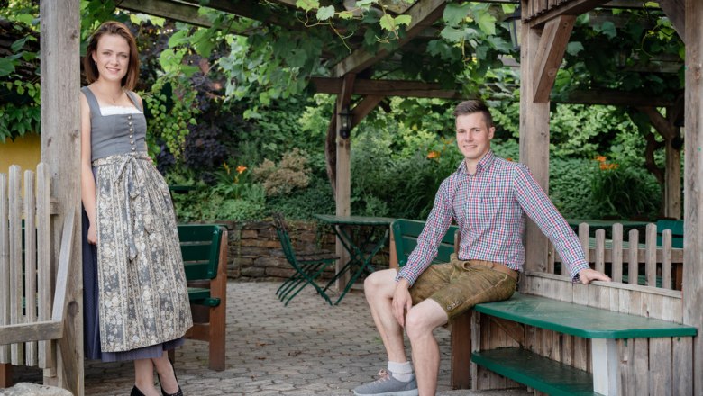 Ein Mann in Lederhose und eine Frau im Dirndl posieren in einem Garten mit Holzkonstruktionen.