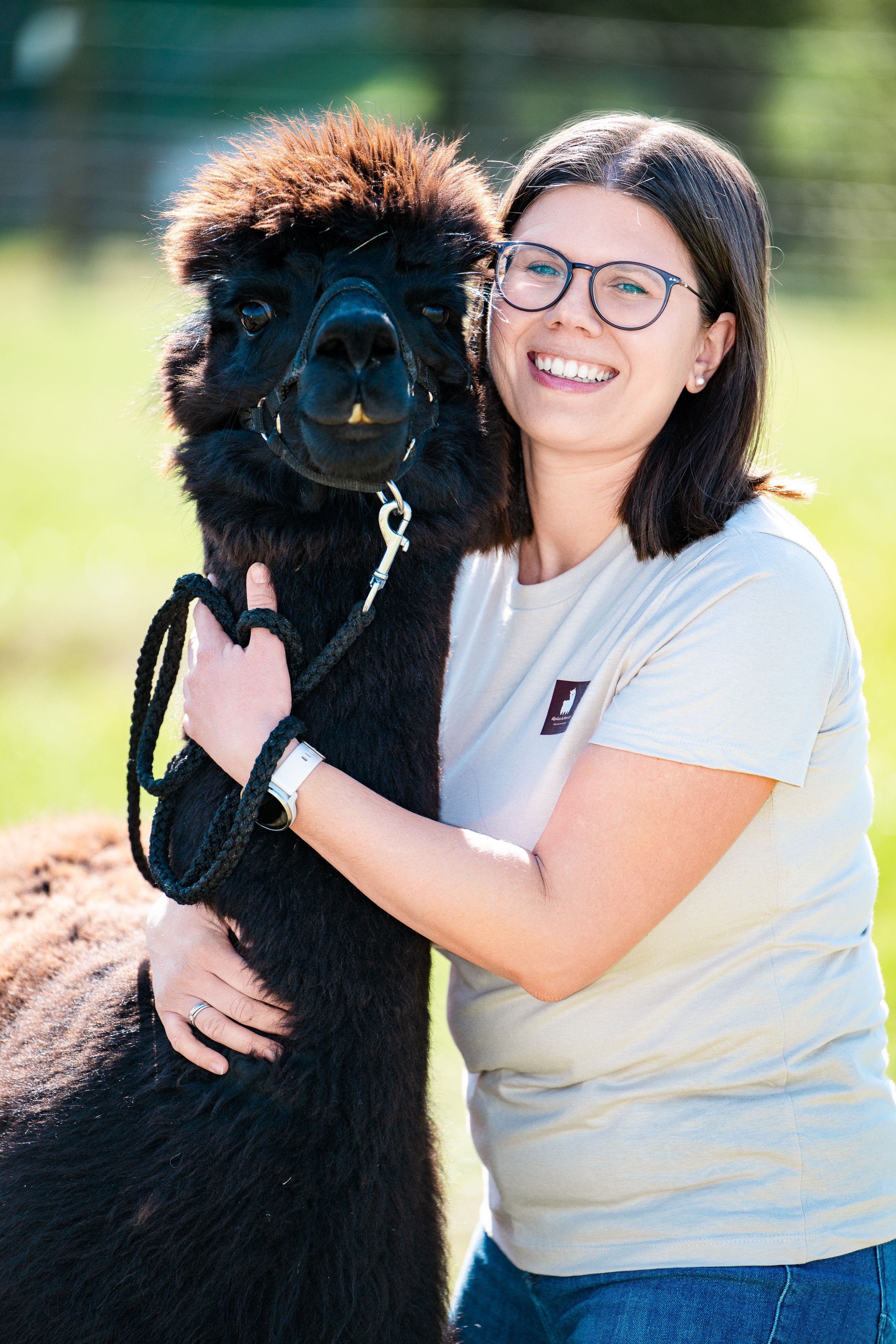 Eine Frau umarmt ein schwarzes Alpaka auf einer Wiese.