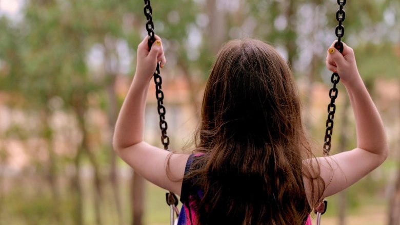 A child swings outdoors, seen from behind, surrounded by trees.