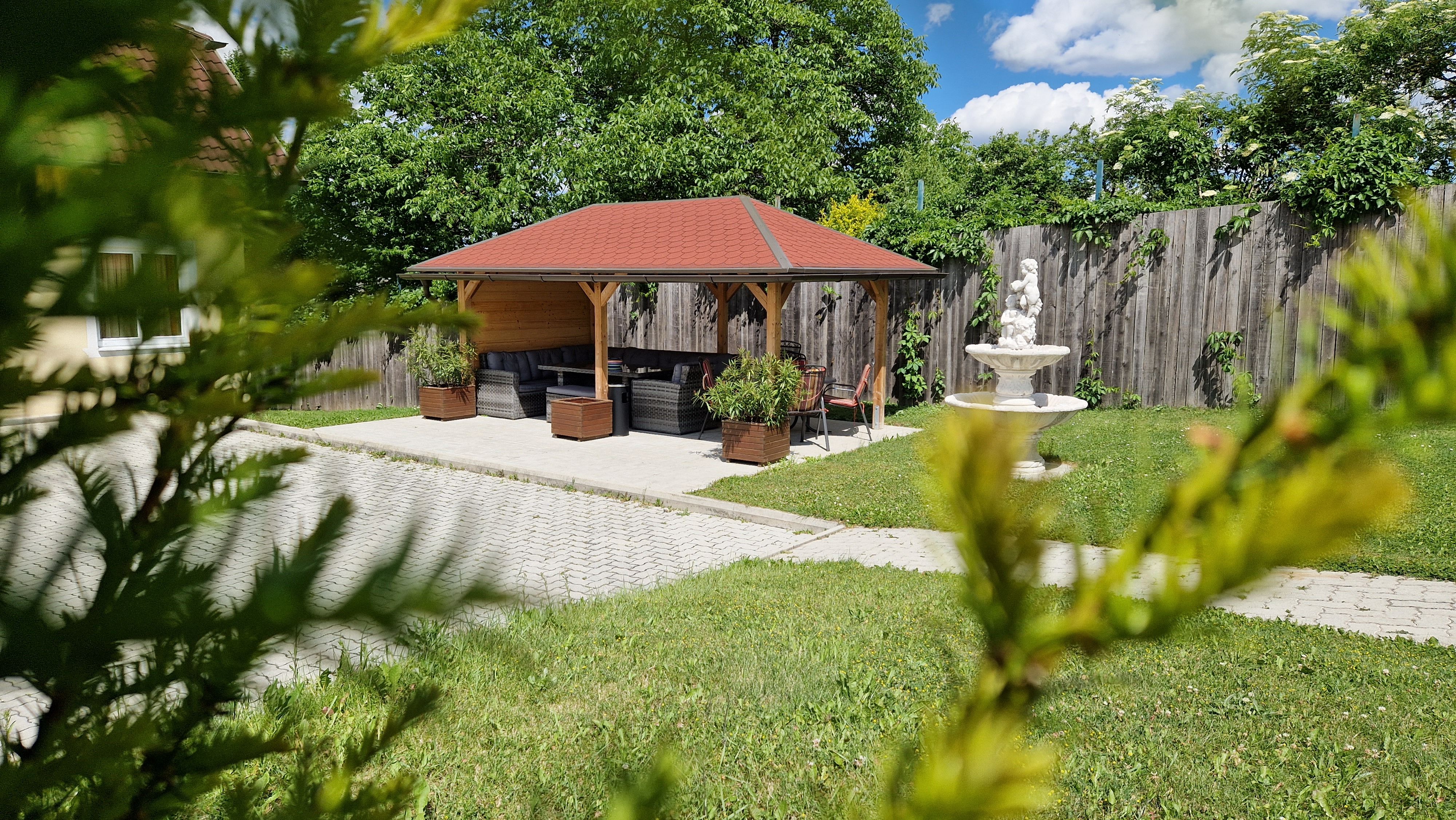 Ein Garten mit Pavillon, Sitzmöbeln und einer Statue auf einem Brunnen, umgeben von Bäumen und einem Holzzaun.