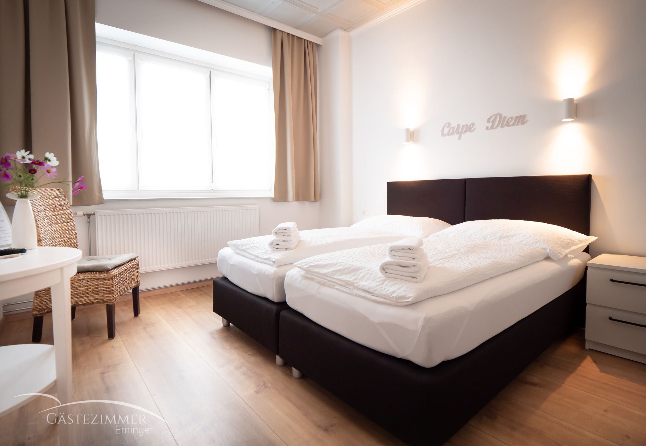 Modernes Doppelzimmer mit zwei Betten, weißen Bettdecken und Handtüchern. Schriftzug 'Carpe Diem' an der Wand, helle Vorhänge und Holzboden.