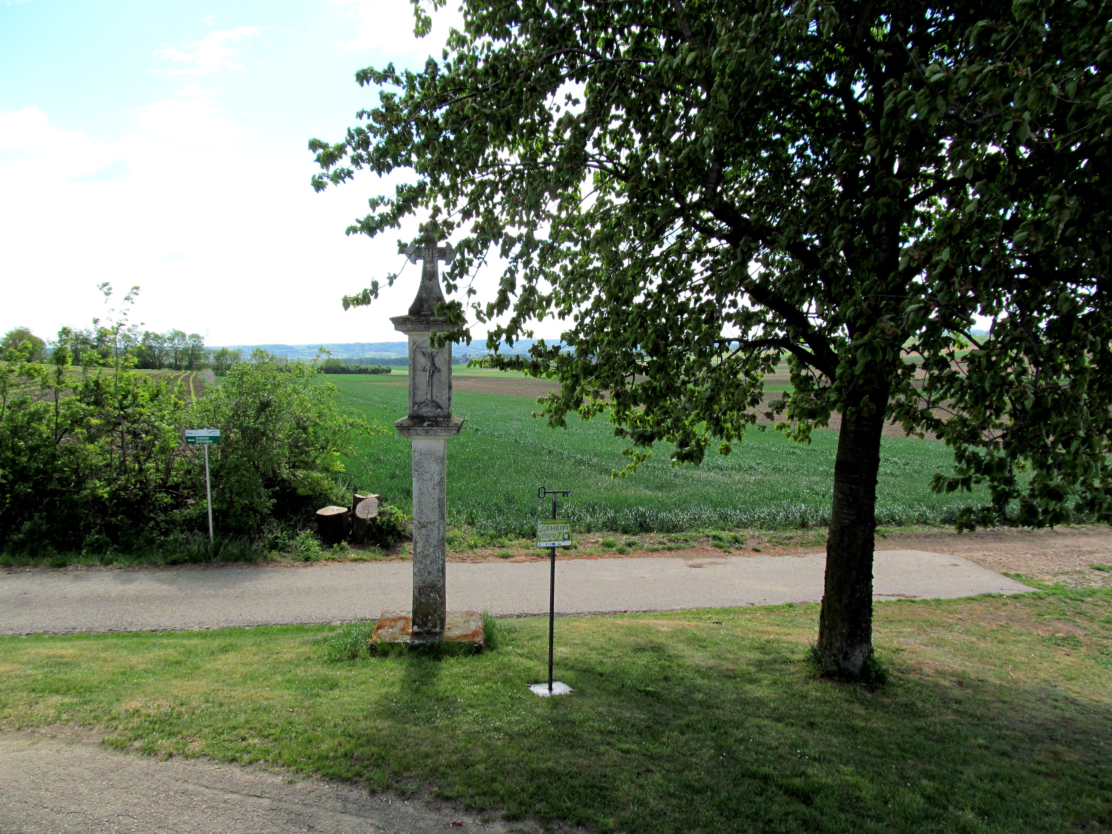 Steinkreuz neben einem Baum auf einer Wiese mit Blick auf Felder.
