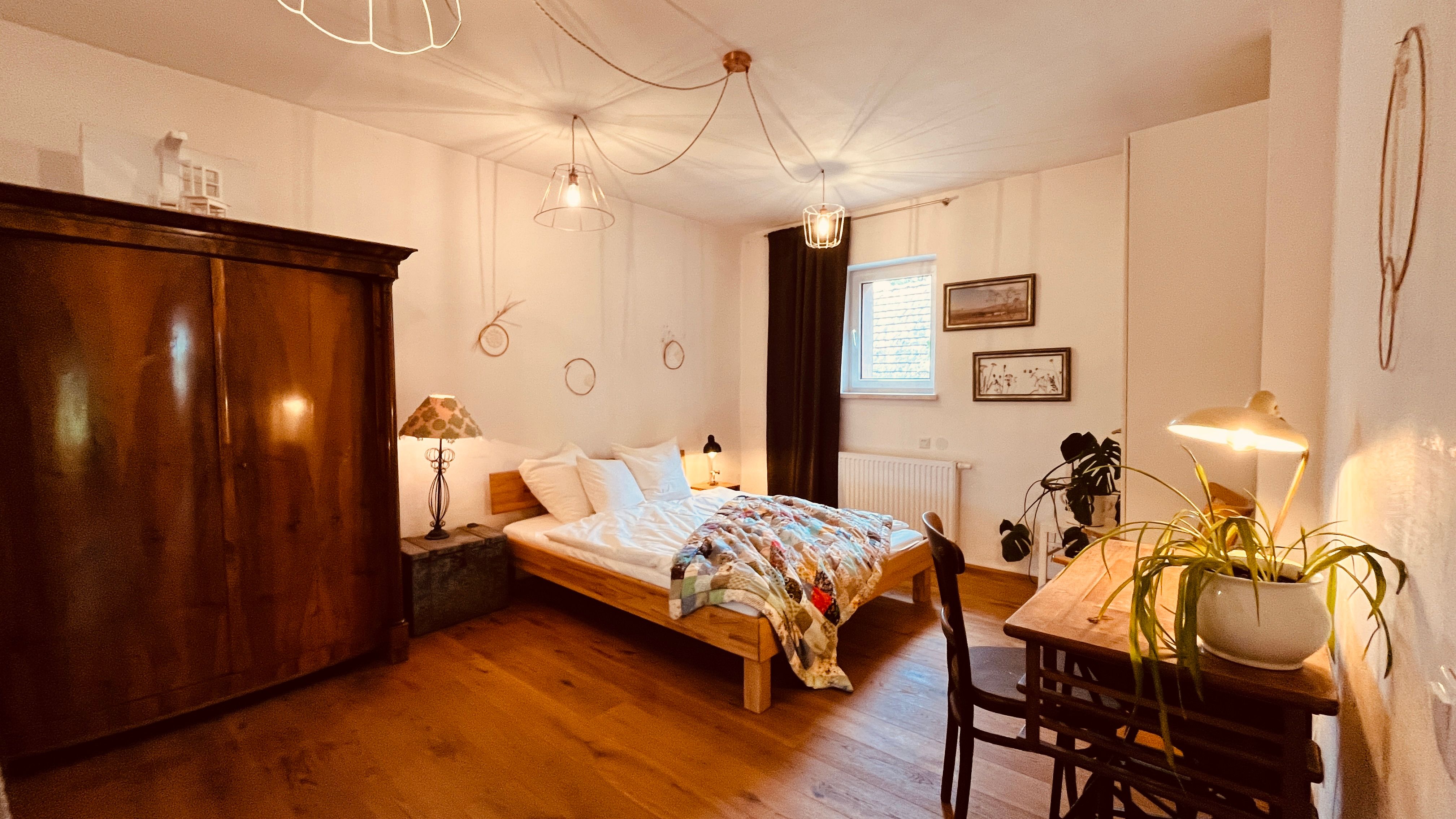 Gemütliches Schlafzimmer mit Holzboden, Doppelbett, antikem Schrank, Schreibtisch mit Lampe und Pflanze, sowie dekorativen Elementen an den Wänden.