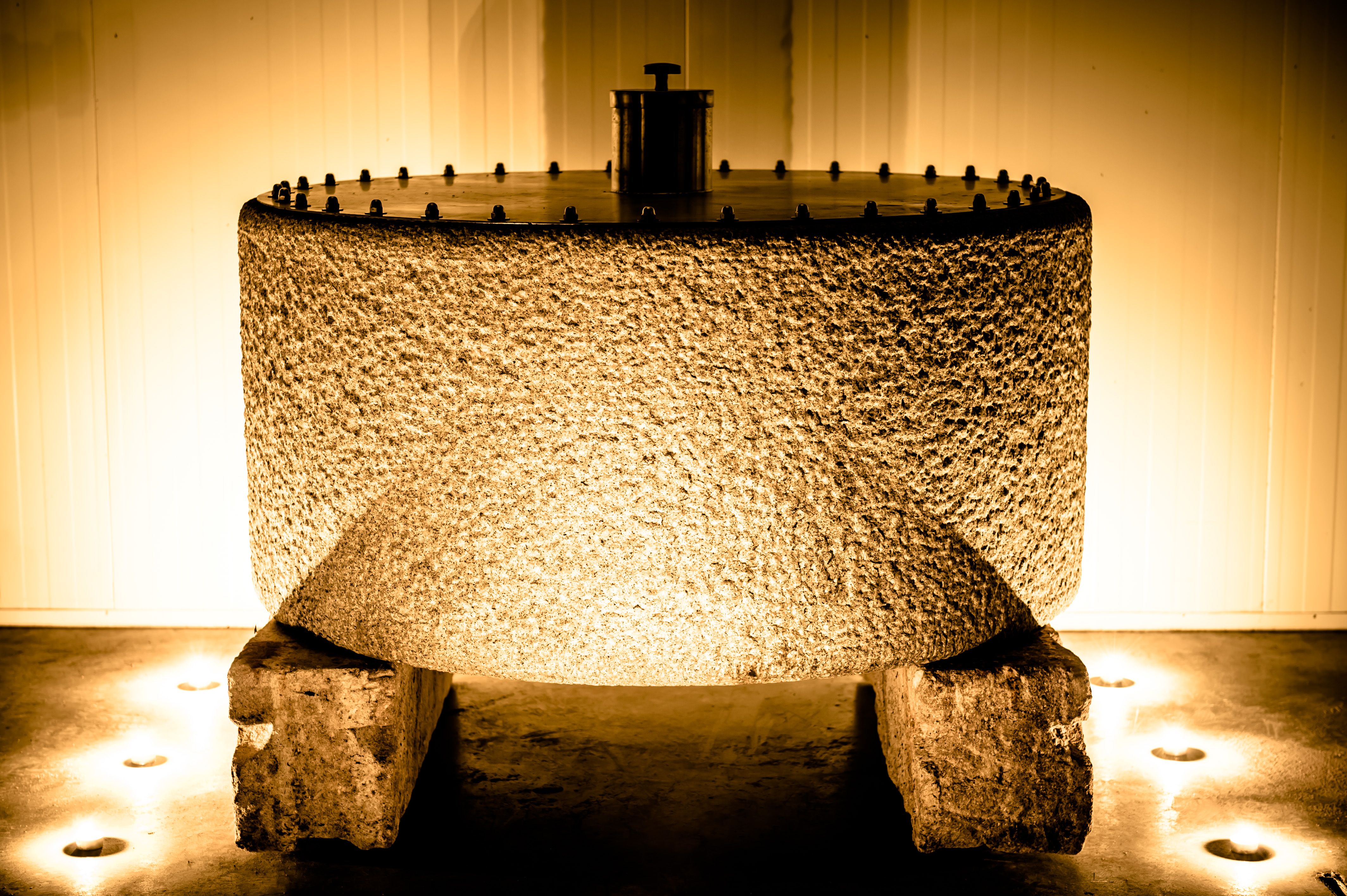 Ein großer, zylindrischer Pressstein aus grobem Material, beleuchtet von warmem Licht, steht auf zwei Steinen in einem Raum.