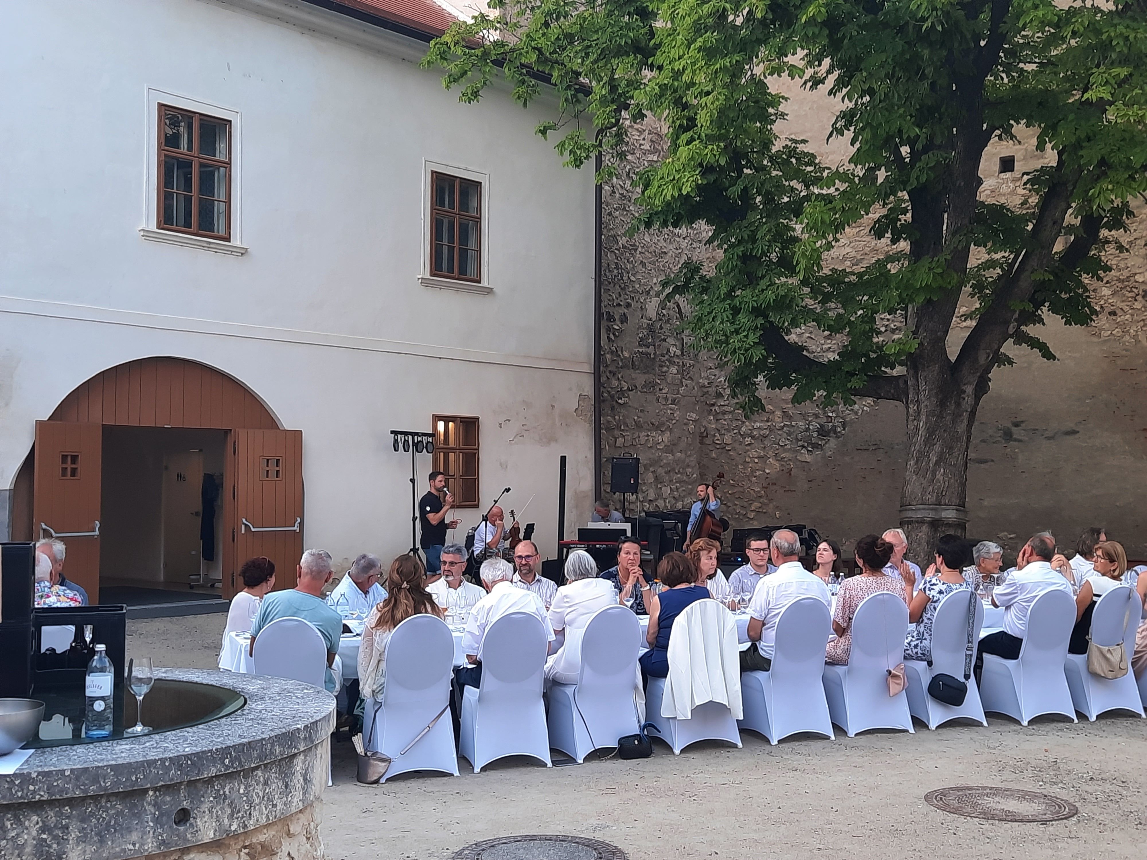 Menschen sitzen an einem langen Tisch im Freien, umgeben von historischen Gebäuden und Bäumen.