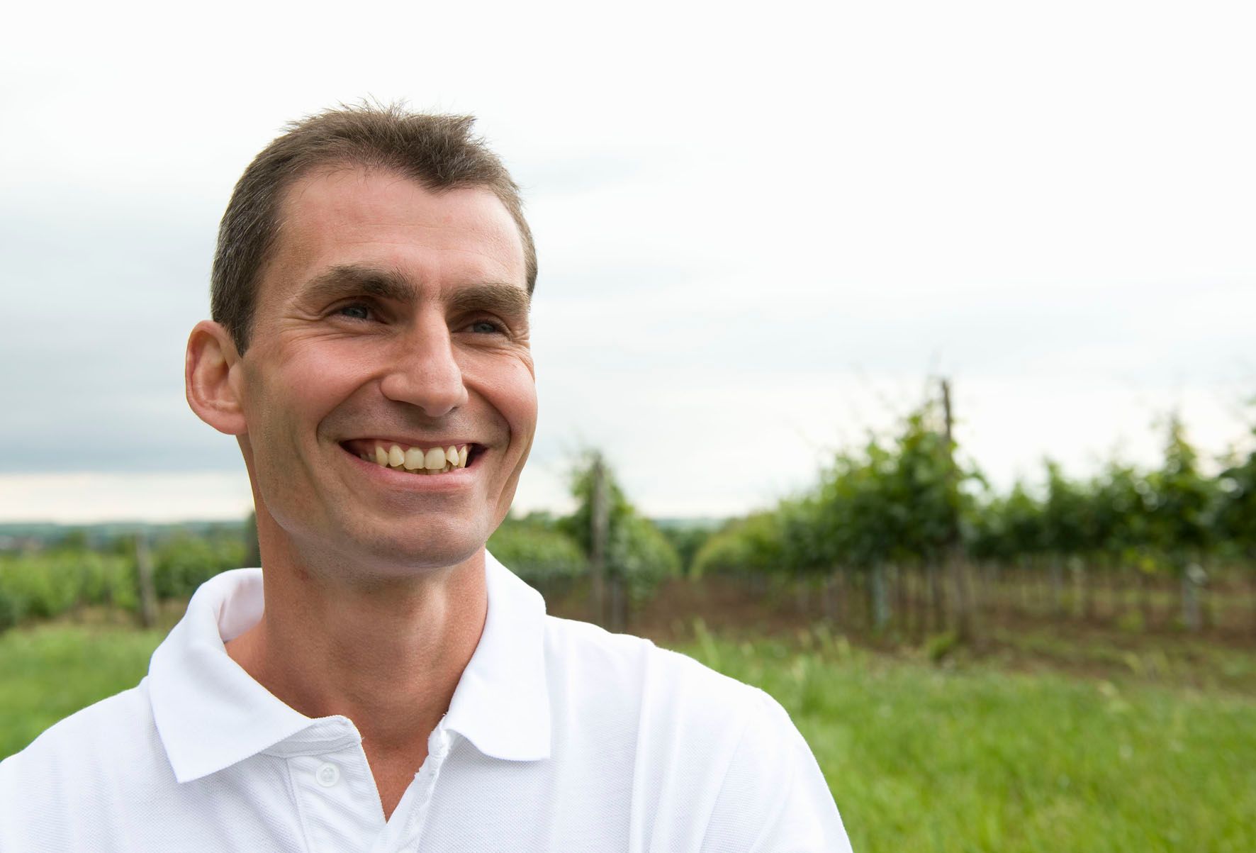 A smiling man in a white polo shirt stands in a field with trees in the background.