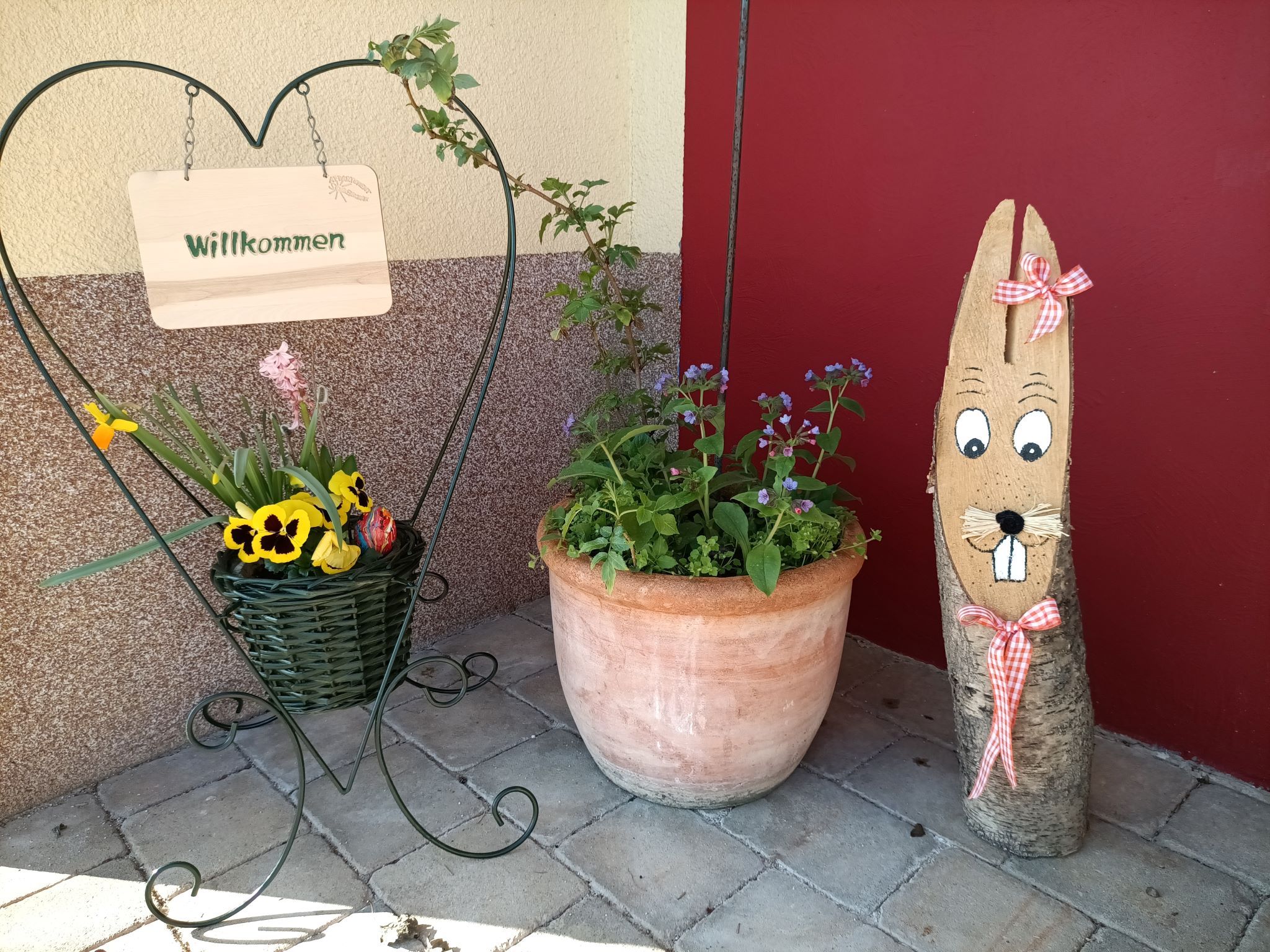 Willkommensschild in Herzform mit Blumen und Holzfigur vor roter Wand.