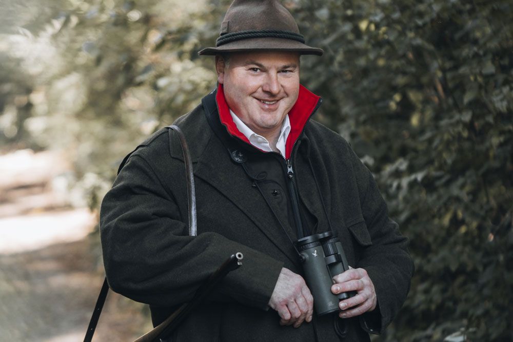 Mann in Jagdkleidung mit Fernglas im Wald.