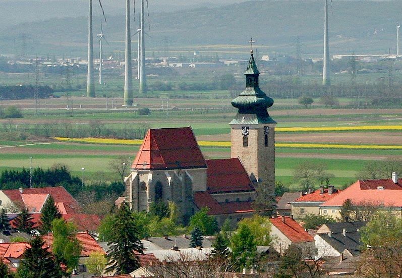 Kirche in Pillichsdorf mit umliegenden Häusern und Windrädern im Hintergrund.