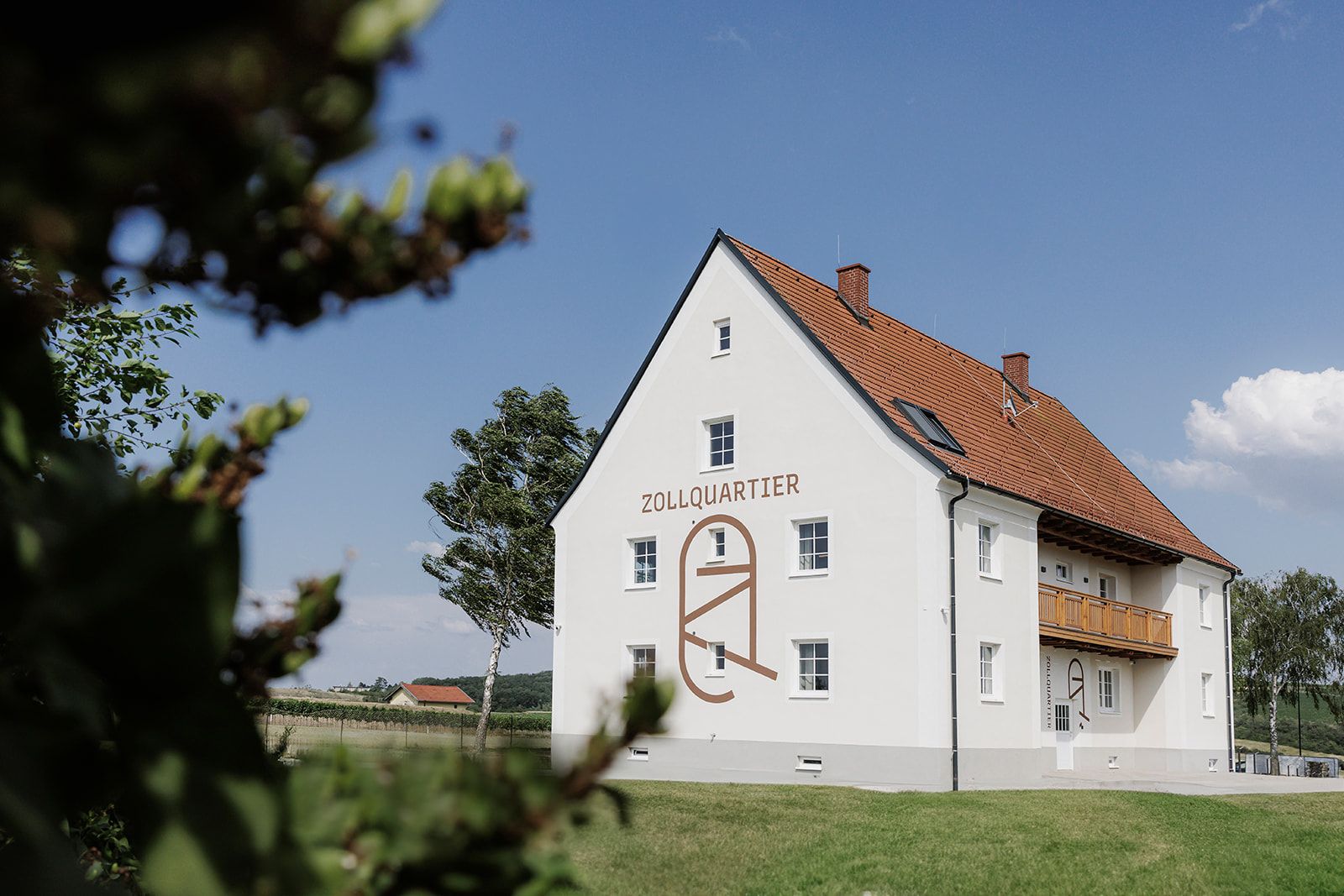 Ein weißes Gebäude mit der Aufschrift 'Zollquartier' und einem roten Ziegeldach in einer ländlichen Umgebung.