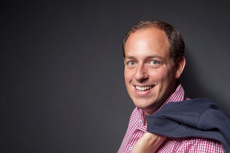 Mann in kariertem Hemd mit Jacke über der Schulter vor grauem Hintergrund.