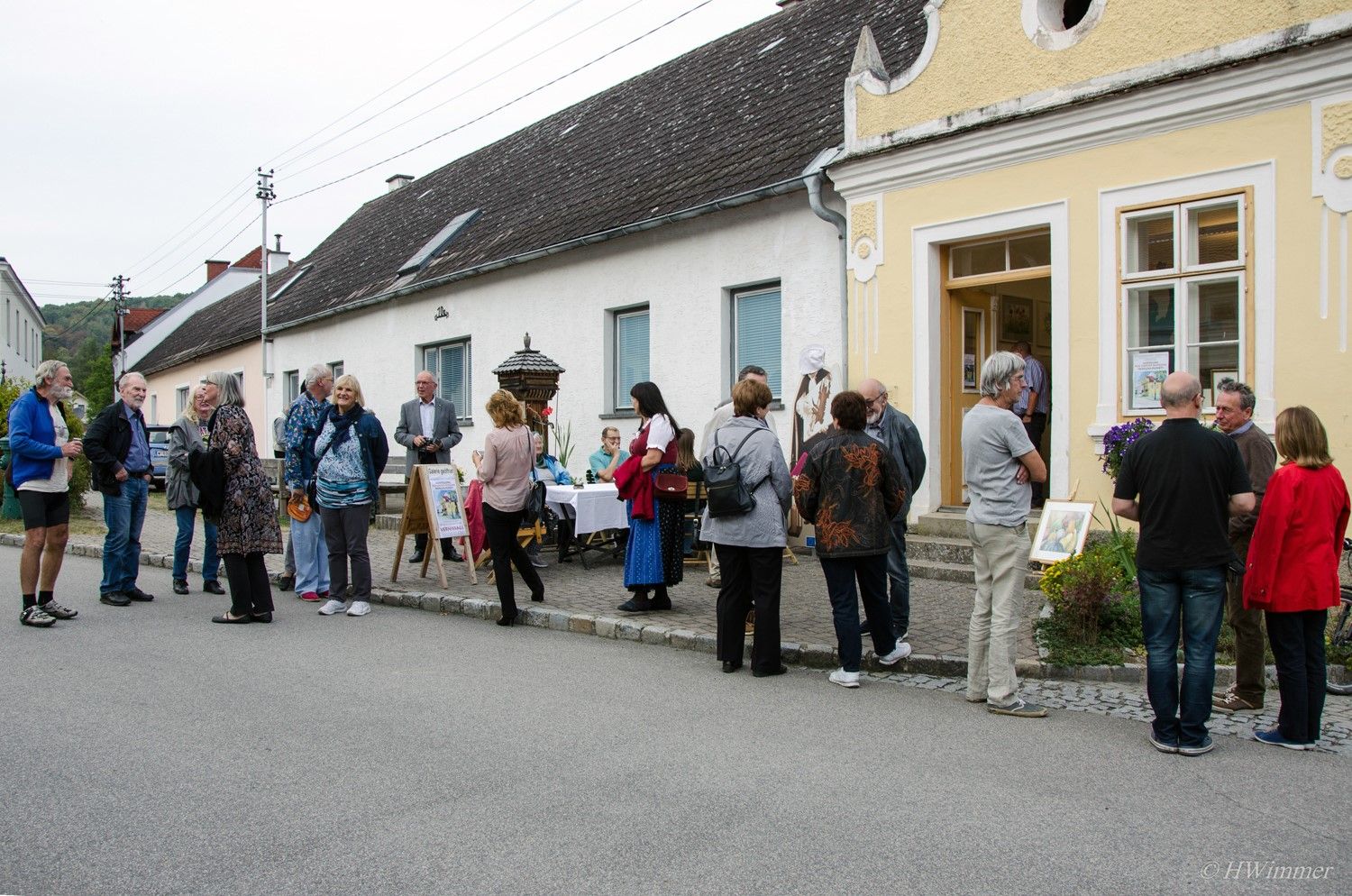 Před žlutou budovou na ulici stojí skupina lidí a povídá si. Někteří z nich si prohlížejí umělecká díla vystavená venku.