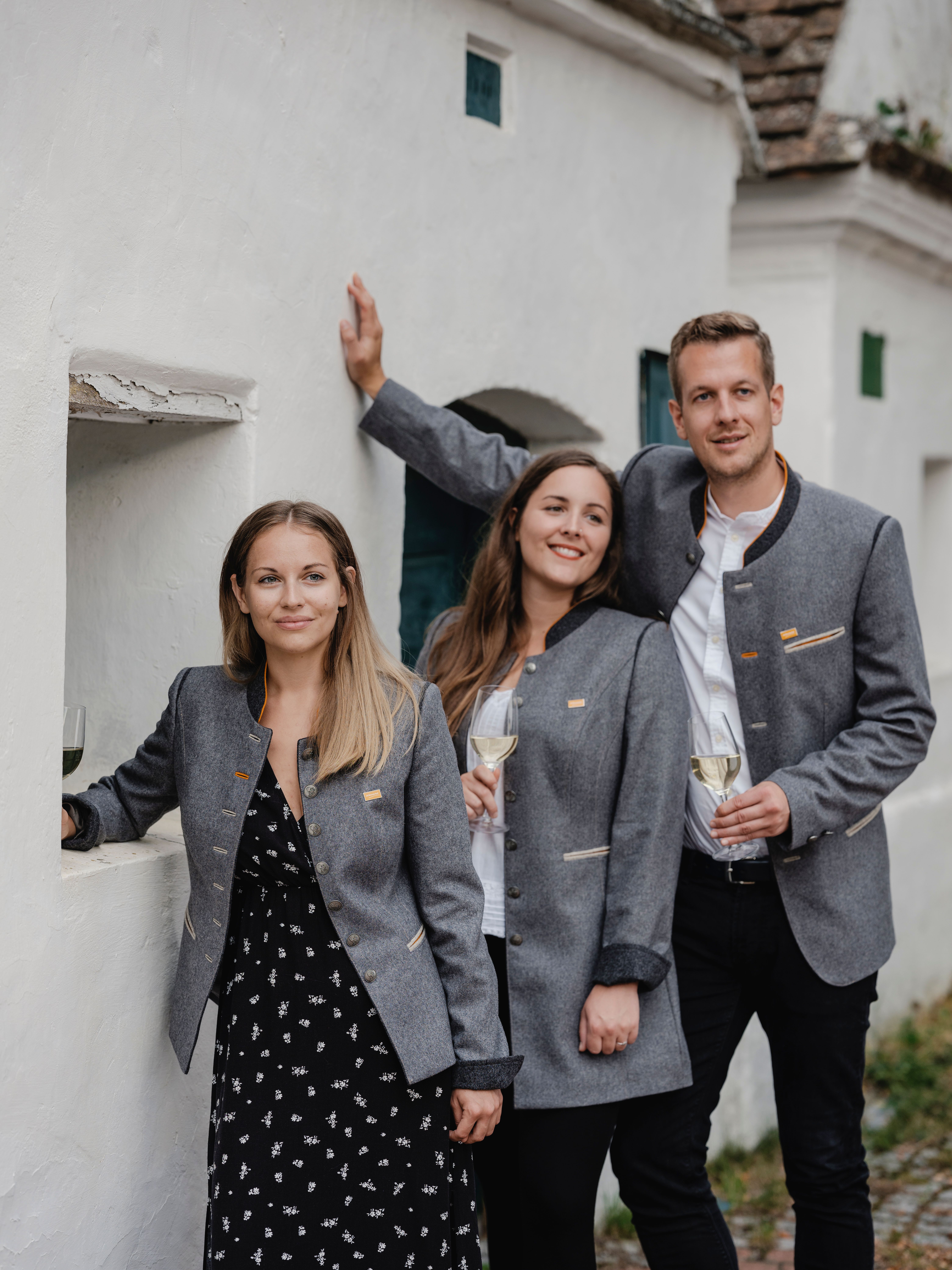 Drei Personen (zwei Frauen und ein Mann) mit grauem Weinviertel-Janker, also in Tracht gekleidet, stehen an die weiß-gekalkte Mauer eines Presshauses in einer Kellergasse gelehnt.