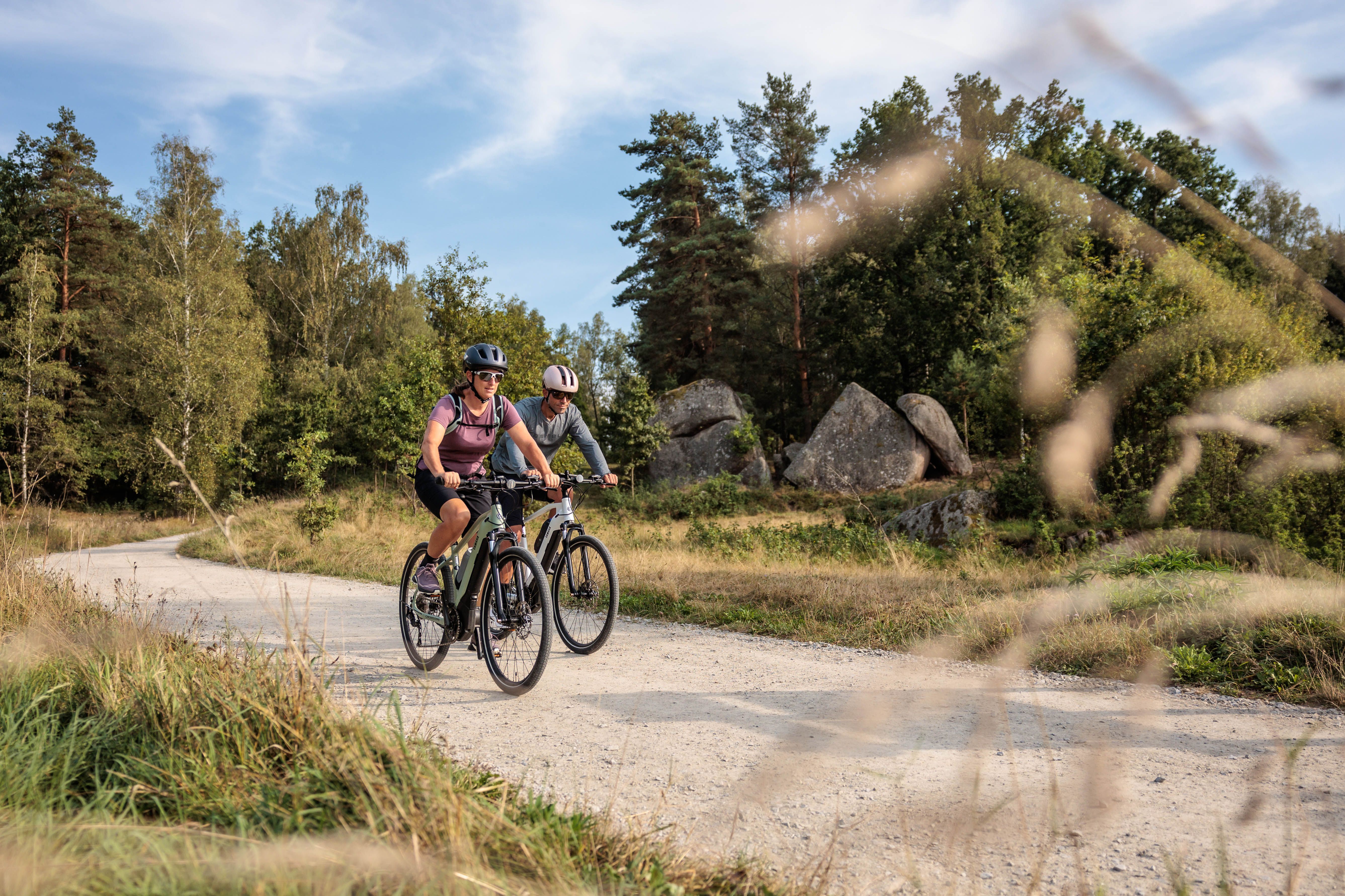 Zwei Radfahrer auf einem Schotterweg im Naturpark Blockheide, umgeben von Bäumen und Felsen.