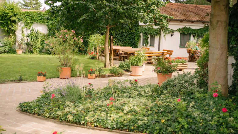 Ein gepflegter Garten mit Rasen, Blumenbeeten und Terrakotta-Töpfen. Im Hintergrund ein Haus mit Terrasse und Holztischen.