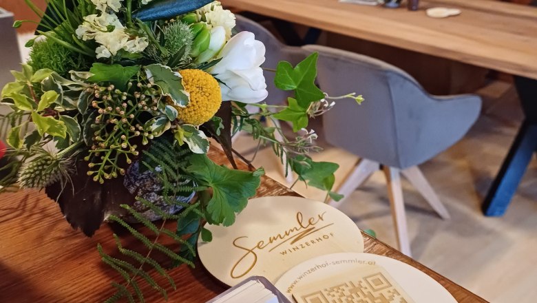 Ein Blumenarrangement auf einem Tisch im Winzerhof Semmler mit QR-Code und Visitenkarten.