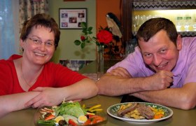 Ein Mann und eine Frau lächeln in die Kamera, vor ihnen stehen Teller mit Essen. Im Hintergrund ist eine rote Rose und ein Regal mit Gläsern zu sehen.